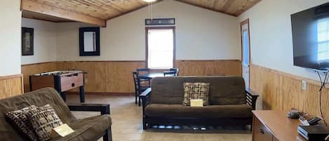 A cozy living room with a wooden ceiling, a sofa, a coffee table, a TV, and a foosball table.