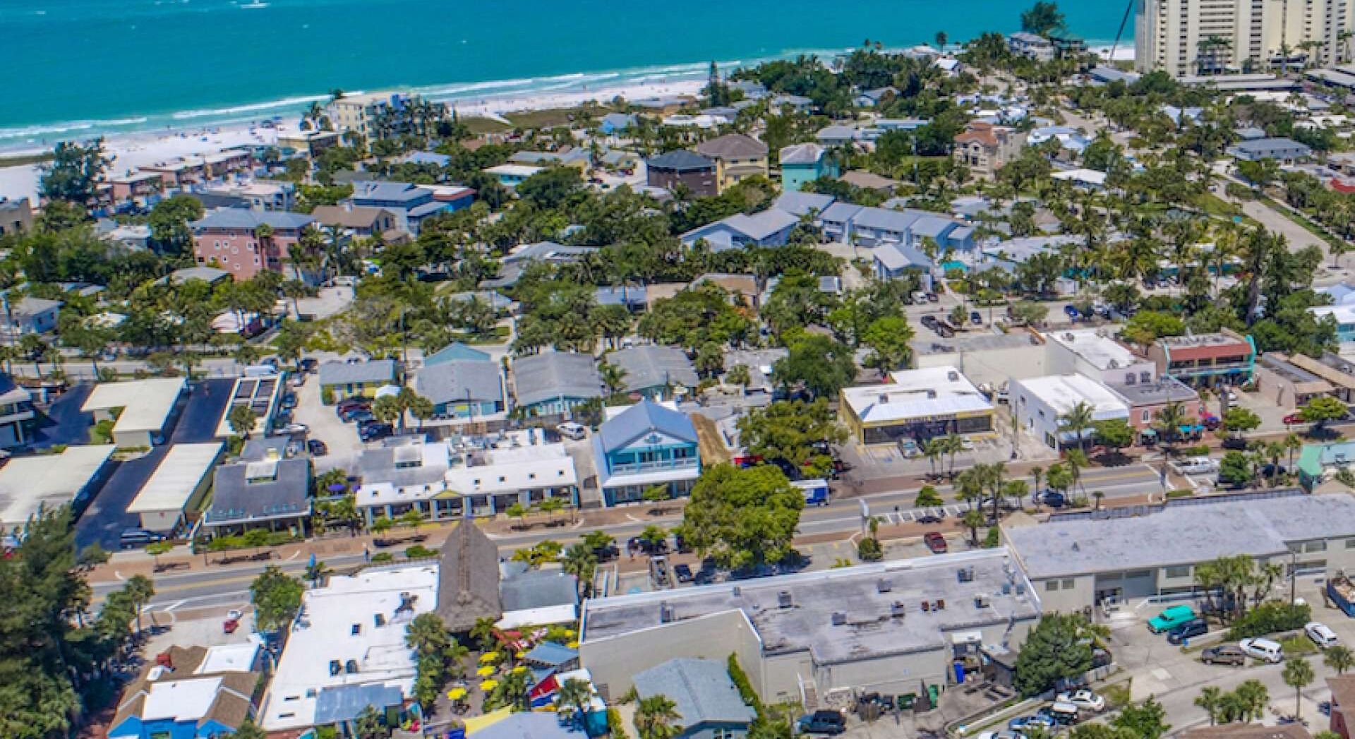 An aerial view of a coastal town with a mix of residential and commercial buildings, a beach, and a clear view of the ocean.
