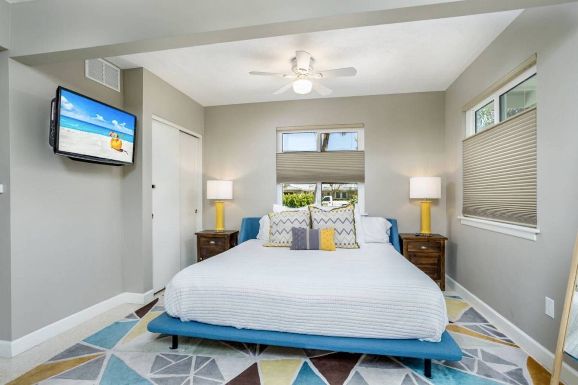 A bedroom with a bed, two bedside tables, a ceiling fan, a TV on the wall, and a window with blinds.