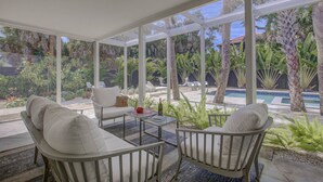A covered outdoor seating area with white cushioned chairs and a glass table, overlooking a pool and lush greenery. - Majestic Coastal Escape (Siesta Key)