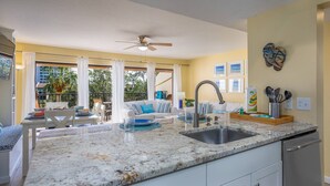 A kitchen with a granite countertop, stainless steel sink, and white cabinets. There is a ceiling fan, a dining area with a table and chairs, and a view of the outdoors through large windows. - Midnight Cove II Bldg 7: Top Floor With Views (Siesta Key)