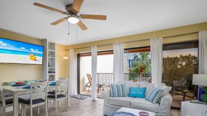 A modern living room with a ceiling fan, a dining area with white furniture, a sofa with blue cushions, and a large window offering a view of the outdoors. - Midnight Cove II Bldg 7: Top Floor With Views (Siesta Key)