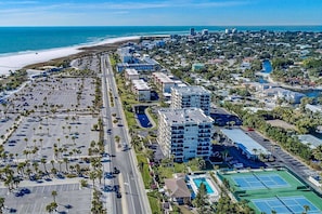 An aerial view of a coastal city with a beach, a large parking lot, and a hotel with multiple floors and a swimming pool.