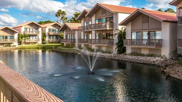 A resort with multiple two-story buildings, each featuring balconies and large windows. A water feature with a fountain is visible in the foreground. - Our House at the Beach Lakeside (Siesta Key)