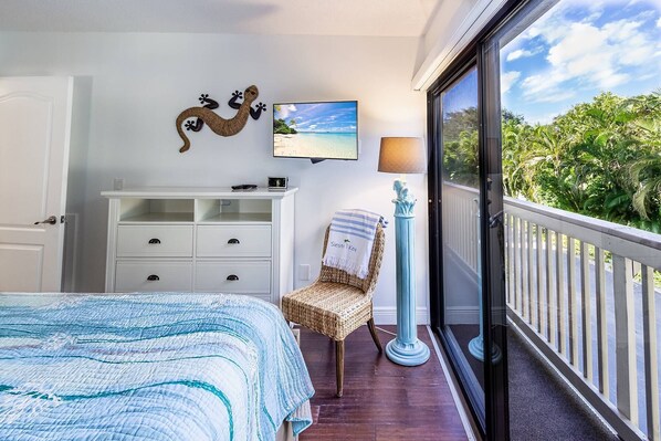 A bedroom with a bed, a dresser, a chair, a TV, and a balcony with a view of greenery.