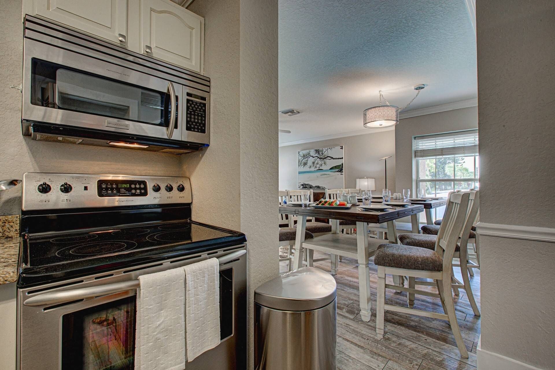 A modern kitchen with a built-in oven, microwave, and stove. There is a dining area with a table and chairs, and a trash can is visible.