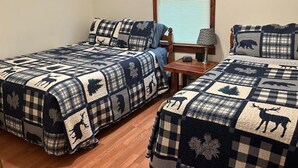 Una habitación con dos camas, cada una cubierta con un edredón a cuadros con estampados de animales. Hay una ventana con persianas, una mesita y una lámpara.