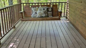 Un porche de madera con un asiento mecededor y cojines, rodeado de árboles.
