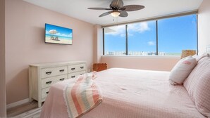 Un dormitorio con una cama grande, ventilador de techo, ventana con vista a la ciudad, cómoda y un cuadro en la pared.