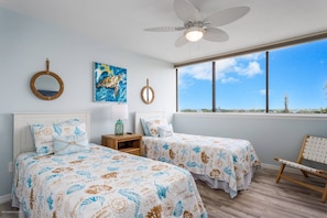A bedroom with two beds, a ceiling fan, a large window with a view, and decorative wall art.