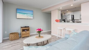 Una sala de estar con sofá, una mesa de centro con flores, televisor y una zona de cocina con refrigerador y taburetes de bar.