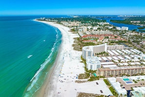 An aerial view of a coastal city with a long sandy beach, a marina, and a mix of residential and commercial buildings.