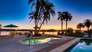 A pool area with a hot tub, lounge chairs, and umbrellas, surrounded by palm trees and a view of a body of water at sunset. - Palm Bay Club - Bayside (Siesta Key)
