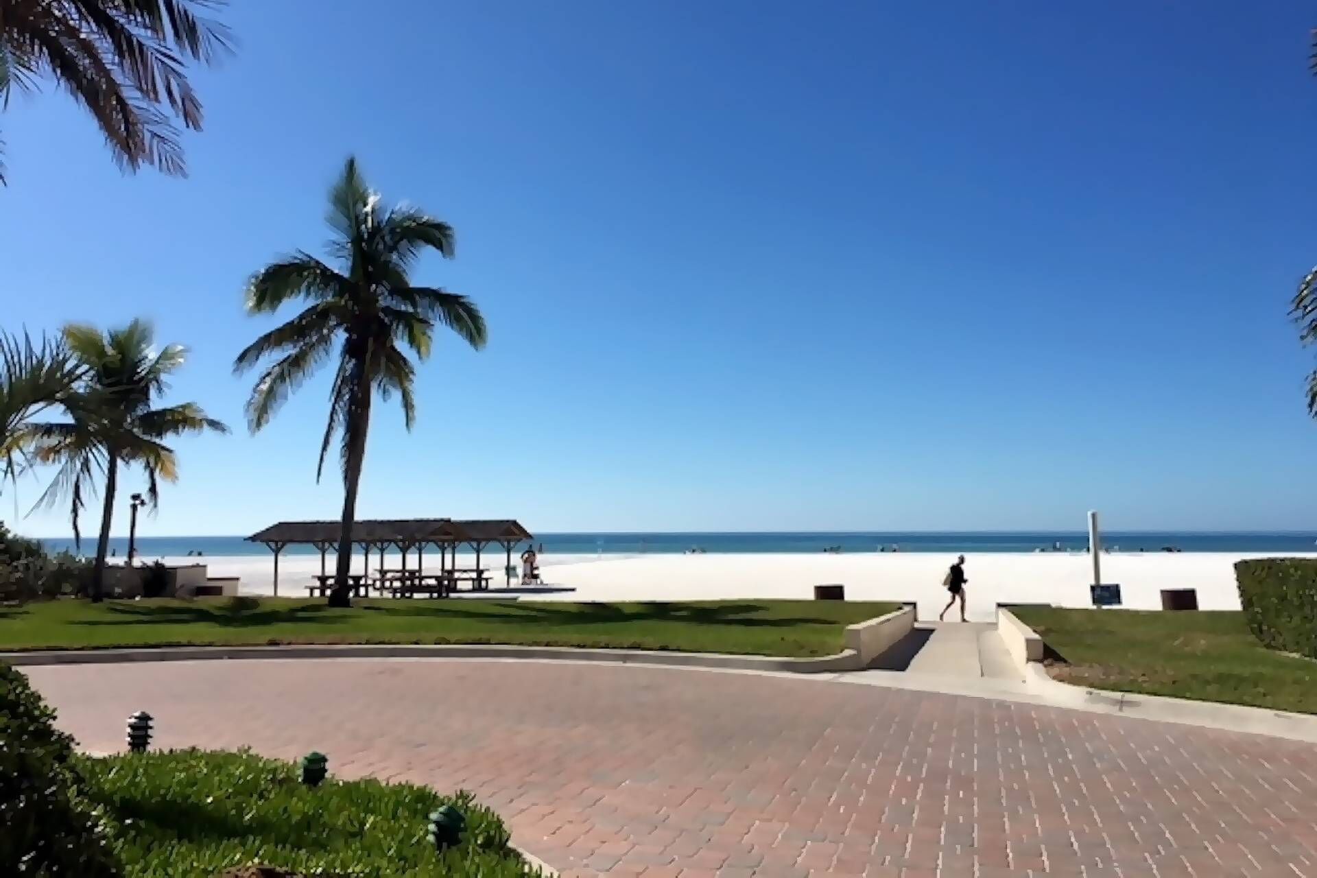 Un sentier pavé mène à une plage bordée de palmiers et dotée d’un pavillon abrité.
