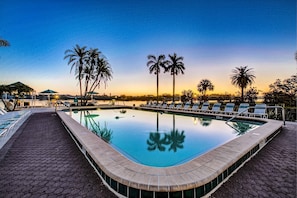 Piscina com espreguiçadeiras e palmeiras, ladeada por um caminho pavimentado.