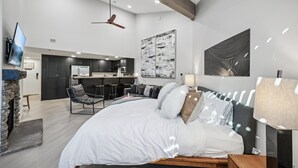 A modern bedroom with a large bed, a fireplace, a dining area, and a kitchen in the background.