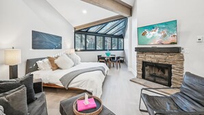 A spacious bedroom with a large bed, a stone fireplace, a dining area with a table and chairs, and a large window providing natural light.