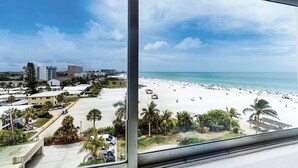 A beach view from a window overlooking a sandy beach with palm trees, a few buildings, and a clear sky. - Palm Bay Club 7th-floor Oceanfront Condo on Private Beach (Siesta Key)