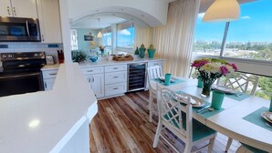 A modern kitchen with white cabinets, a microwave, and a stove. There is a dining area with a table and chairs, and a view of the city through large windows. - Palm Bay Club 7th-floor Oceanfront Condo on Private Beach (Siesta Key)