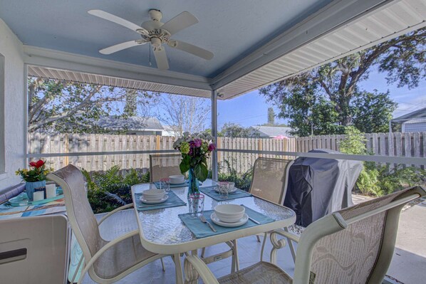 Une terrasse couverte avec une table dressée pour deux personnes, des chaises, un ventilateur de plafond et une vue sur une clôture en bois et des arbres.