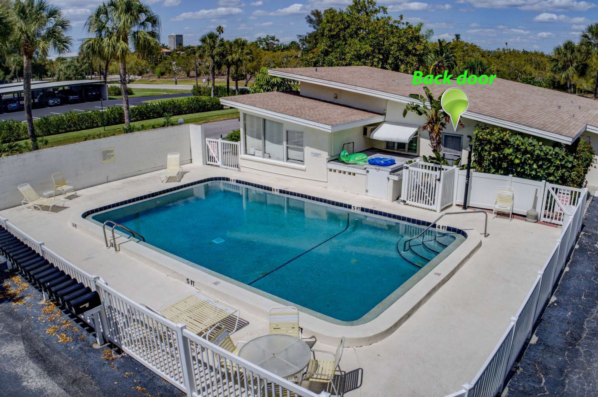 A rectangular swimming pool with a white deck, surrounded by a white fence. There are lounge chairs and a small table near the pool. A building with a red roof is visible in the background.