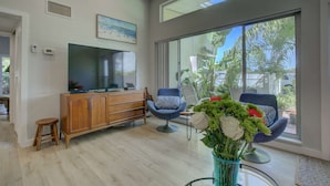 Ein modernes Wohnzimmer mit Flachbildfernseher, Holzschrank, zwei blauen Sesseln und einem großen Fenster mit Blick auf Grünflächen.