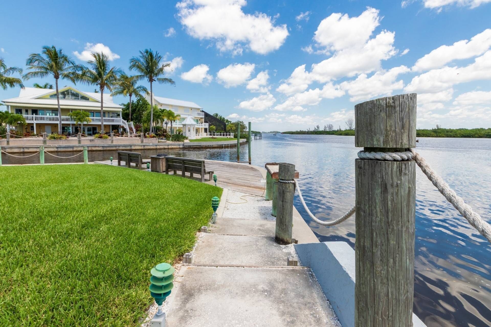 A waterfront walkway with a wooden post and rope barrier, a grassy area, and a building with a white roof and balconies.