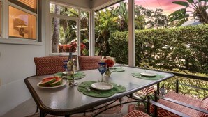 Un coin repas avec une table dressée pour deux personnes, un vase rempli de fleurs et une vue sur une végétation luxuriante et un coucher de soleil.