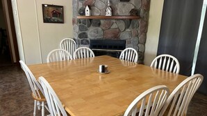 Un comedor con una mesa de madera y sillas blancas, una chimenea de piedra y un pequeño estante con adornos.