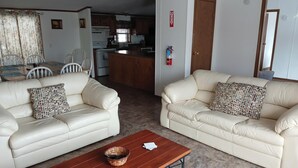 Una sala de estar con dos sofás color crema, una mesa de centro de madera con revistas, y una cocina al fondo.