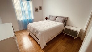 Une chambre à coucher comprenant un lit, des tables de chevet, une commode, une fenêtre avec des rideaux et un mur orné de deux tableaux encadrés.