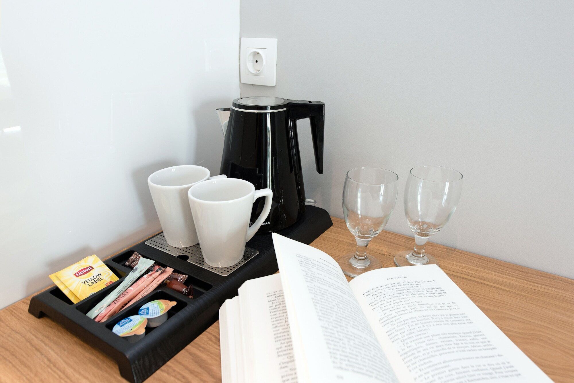 Une machine à café, deux tasses blanches, deux verres à vin, un livre et un plateau contenant divers objets, le tout posé sur une table en bois.