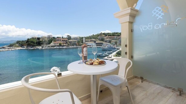 A balcony with a table set for two, overlooking a marina and coastal town. - Melina Bay Boutique Hotel (Corfu)