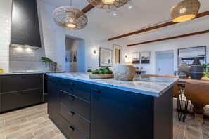 Une cuisine moderne dotée d’un plan de travail en marbre, de meubles de rangement aux couleurs sombres et d’un grand îlot central. On y trouve des luminaires suspendus en osier, un coin repas avec une table en bois et divers appareils de cuisine.