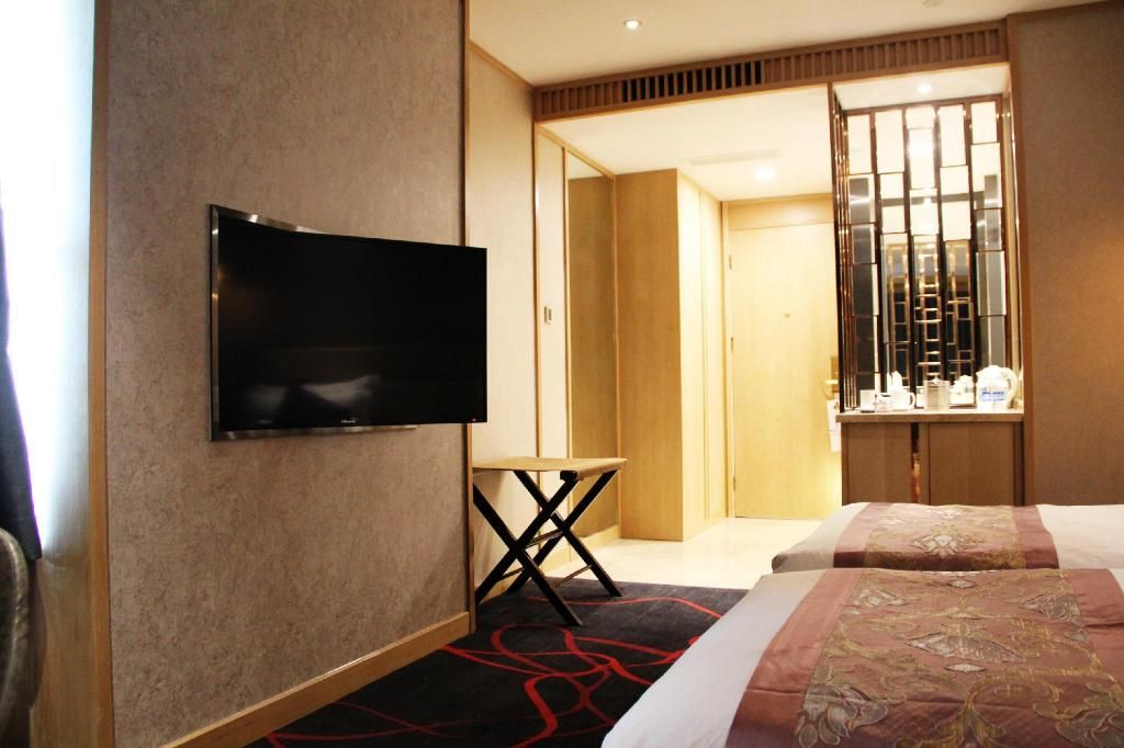 A hotel room with a flat-screen TV mounted on the wall, a small table, a bed with a patterned bedspread, and a wooden wardrobe.