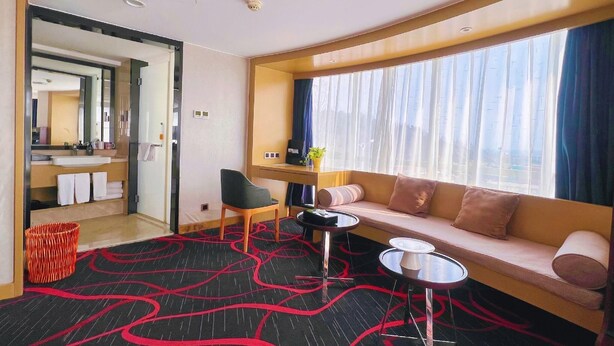 Una habitación de hotel moderna con sofá, una mesita, una silla y un gran ventanal con cortinas.
