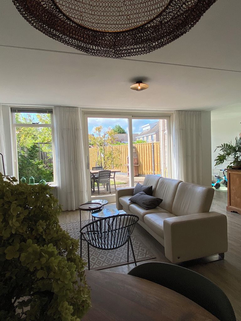 A modern living room with a sofa, coffee table, and chairs. There is a large window with curtains, a balcony, and a view of the outside.