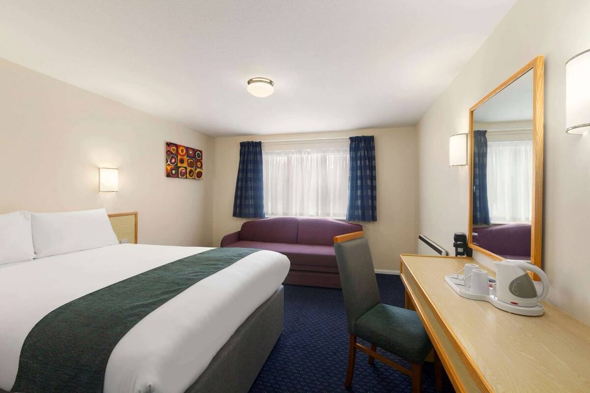 A hotel room with a bed, a sofa, a desk with a chair, a coffee maker, and a mirror.
