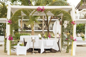 A beachside outdoor dining area with a white table set for two, surrounded by palm fronds and pink flowers.