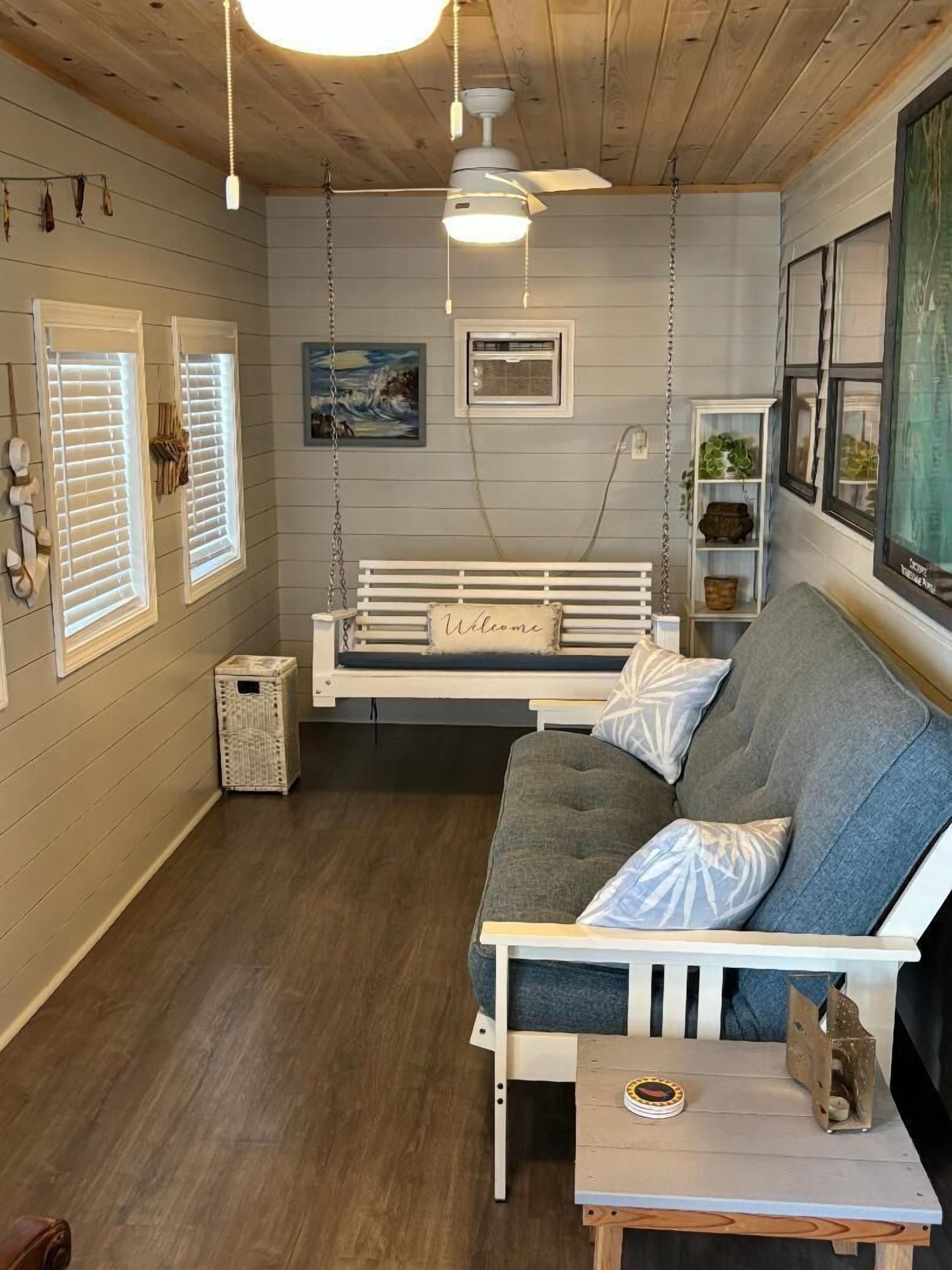Une pièce chaleureuse avec un plafond en bois, un banc blanc orné d’une pancarte « Bienvenue », un canapé gris, une petite table sur laquelle est posé un objet décoratif, et une étagère murale agrémentée de plantes.