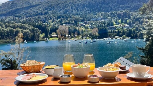 Una mesa de desayuno con diversos alimentos y bebidas, con vistas a un lago con barcos y un paisaje montañoso.