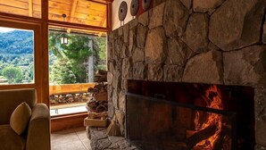 Una chimenea de piedra con fuego, un banco de madera y vistas de un paisaje boscoso a través de amplios ventanales.