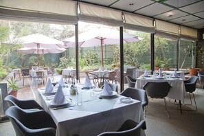Un área de comedor al aire libre con mesas preparadas para dos, cubierta por sombrillas y rodeada de follaje.
