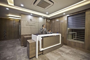 A modern reception desk with a man standing behind it, a clock on the wall, and a large window with blinds.