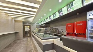 A modern food counter with a glass display case, a Coca-Cola refrigerator, and a menu board above. - Hotel Sifat International (Surat)