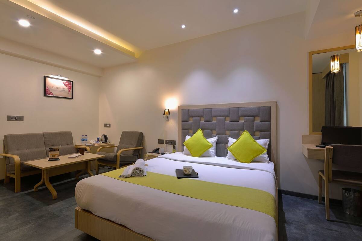 A hotel room with a bed, two armchairs, a small table, a desk, a mirror, and a wall-mounted lamp.