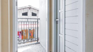 Eine geöffnete Tür, die auf einen Balkon mit Metallgeländer, einen gefliesten Boden und einen Blick auf ein Gebäude mit Wellblechdach führt.
