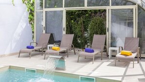 Un espace aménagé au bord de la piscine, avec des chaises longues, un élément décoratif aquatique et un espace clos par des parois de verre, agrémenté de verdure.