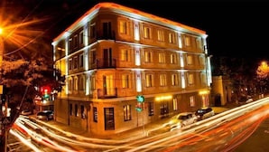 Un immeuble à plusieurs étages, doté de fenêtres et de balcons illuminés, entouré d’une rue animée où l’on aperçoit des traînées de lumière laissées par les véhicules en mouvement.