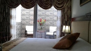 Una habitación de hotel con cama, ventana con cortinas, balcón con sillas y un florero con flores.